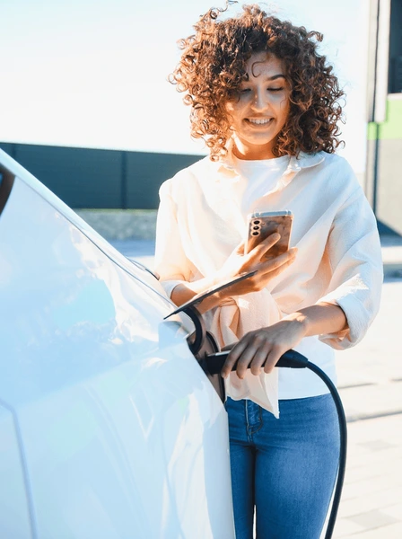 Afbeelding van vrouw die elektrische auto oplaadt en een app op haar telefoon bekijkt.
